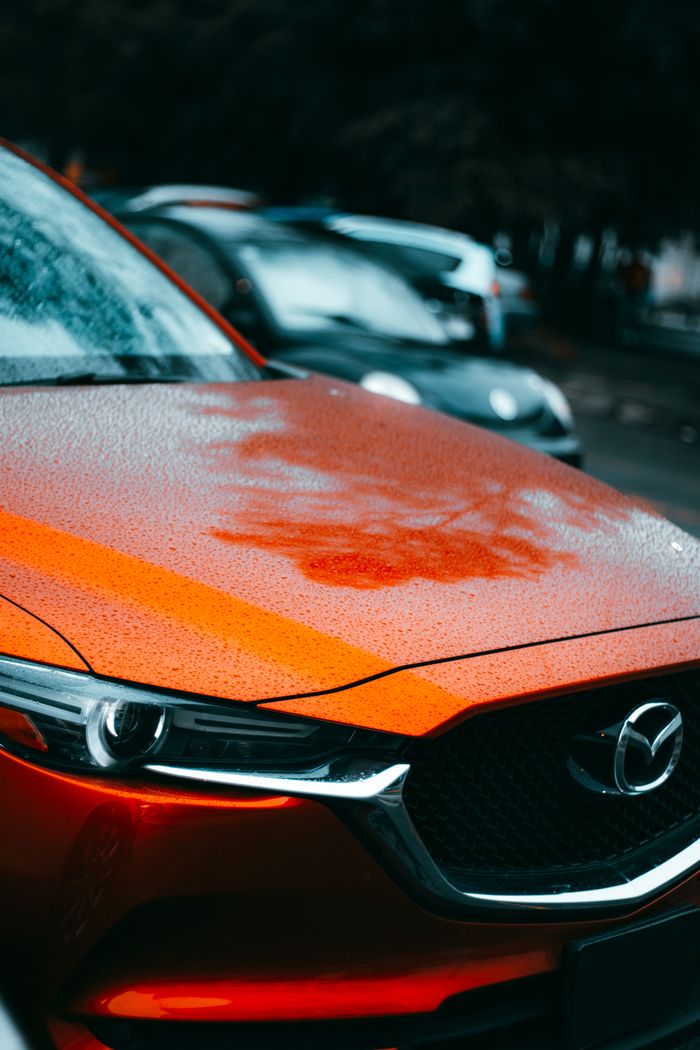team-03 Detailed view of a red car's bonnet with raindrops, showcasing its sleek design and vibrant color in a rainy outdoor setting.